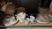 White ceramic kitchenware including teapot, creamer set, mugs, plates, soup tureen, and clear glass divided serving dish