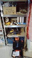 Shelf with various hardware parts and tools including extension cords, hardware pieces in labeled boxes, hand tools and small hardware items.