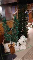 View of two faux trees with artificial green ivy leaves in white distressed wooden planters. A small white plastic ghost figure is placed to the right and a plush moose head with decorated antlers is at the front on the floor.