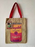 Front view of beige canvas tote bag with red straps, showing silkscreened multicolor Campbell's Tomato Soup can design and attached kraft hangtag with three pins.