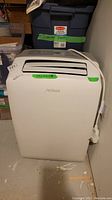 Front view of white Noma portable air conditioner with vents and control panel on top, placed on basement floor with a green tape label.