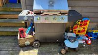 Front view of CharBroil propane BBQ grill with 4 burners and side burner, showing control knobs and temperature gauge on lid, set against an outdoor wooden deck with toys nearby.
