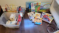 Overview of all items in the lot including books, toys, and craft items arranged on the floor and in the plastic bin.