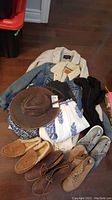 Photo of folded clothing items including Levi's sherpa jacket and Andrew Mark trench jacket, a leather brimmed hat by Goorin bros, patterned dresses, black thigh-high boots, L.L. Bean moccasins, and casual shoes and boots. All items laid on brown wooden floor.