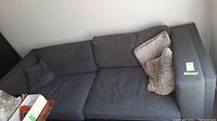 View of the large dark gray Ikea couch with two small dark gray throw pillows and one larger light brown fuzzy throw pillow on the right armrest, showing its six seat and back cushions and wooden legs.