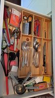 Drawer partially filled with metal forks, spoons, a small box grater, red-handled scissors, a few other utensils, and a long knife with black handle