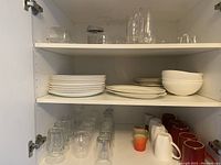 Shelf displaying stacks of plates including diner and side plates, ceramic bowls, glassware, and cups.
