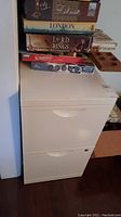 Front view of beige two-drawer metal filing cabinet with several board games stacked on top, bottom drawer slightly open showing hanging folders.