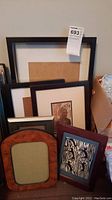 Five picture frames of varying styles and colors leaning against a wall. Includes wooden and plastic frames. One frame shows lithograph carving artwork.