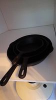 Three black cast iron frying pans stacked by size on white shelf