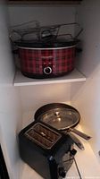 Red plaid Crock Pot slow cooker on shelf with metal cooking rack on top. Frying pan with glass lid and black Cuisinart toaster below.
