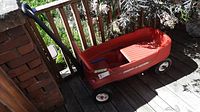 Front and side view of red Radio Flyer wagon placed on wooden deck showing handle, seats, and wheels including taped object on back left wheel.