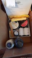 Top view of UGG shoe care kit contents including conditioner, shoe renew, protection sprays, mink oil tin, shoe polish tin, two brushes, and cleaning cloths inside a cardboard box