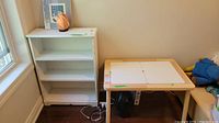Photo showing white wood bookcase with three shelves and kids table beside it