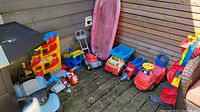 View showing all outdoor kids toys arranged on wooden floor against wooden wall, including ride-on vehicle, multi-level car track, various toy vehicles, push mower, and sled