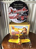 Photo showing the round Coca-Cola metal sign on top featuring an airplane with 'Your thirst takes wings' text, the yellow die-cast airplane coin bank model on the table beneath, and the box for the coin bank behind the airplane.