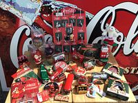 Wide shot of various miniature Coca-Cola themed Christmas collectibles arranged on a table with Coca-Cola sign background