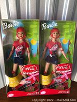 Two Coca-Cola Barbie dolls in clear-front boxes showcasing the dolls dressed in branded t-shirts, shorts, helmets, and including skateboards.