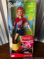 Front boxed view of Coca-Cola Barbie doll with helmet, red shirt, denim shorts and included skateboard.