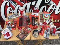 Photo showing entire lot of Coca-Cola branded collector items arranged on a wooden surface including toys, plush, pens, keychains, and packaging with large Coca-Cola wall sign backdrop.