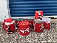 Photo of all 7 Coca-Cola metal containers grouped outdoors showing various types and sizes including large insulated jug, buckets, canisters and cooler style containers.