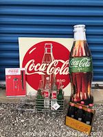 Full view of all items: metal Coca-Cola sign, large bottle display, wooden crate with bottles, metal bottle carrier, and Coca-Cola calendar booklet.
