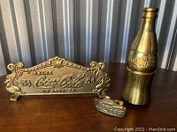 Set of three yellow brass Coca-Cola items including bottle figurine, vintage-style sign, and bottle opener on wooden surface with metal backdrop