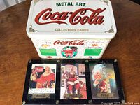 White metal tin box with Coca-Cola logo and three collector cards displayed on the lid showing Santa Claus artwork.