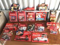 Image showing the full collection of 18 Coca-Cola model vehicles, some in original packaging and some loose, arranged on a shelf with visible logos and colors.