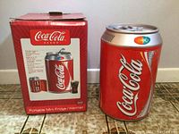 Portable Coca-Cola branded mini fridge or warmer next to original box on tiled floor.