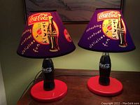 Pair of Coca-Cola table lamps, purple shades with colorful Coca-Cola logos and bottle designs, red bases, shown on wooden surface.
