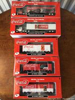 Six Coca-Cola Train Collection Series A model vehicles including two Kenworth W-45 trailer trucks and four 40-foot steel reefer train cars, all in original red Coca-Cola packaging arranged on a table.