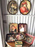 Full view of eight vintage metal trays displayed on a wooden table and metal wall surface, showing artwork of women in period attire and various Coca-Cola and Pepsi-Cola branding.