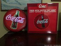 Photo showing the Coca-Cola fiber optic bottle cap lamp lit with multi-colored lights next to its original box with branding and lamp image.