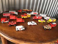 Full view of 14 Coca-Cola model vehicles on wooden table, various styles and sizes including trucks, vans, race car, jeep, and boat trailer.