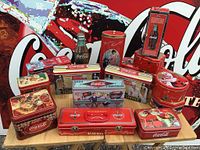 Front view of entire collection of Coca-Cola tins with assorted shapes and vintage designs, displayed against a Coca-Cola sign background.