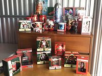 Group photo showing Coca-Cola holiday collectibles including ornaments, boxed decorations, and plush toys arranged on wood furniture.