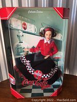 Front view of Barbie doll in original box. Doll dressed in red sweater and black checkered Coca-Cola skirt, visible accessories include a small Coca-Cola bottle.
