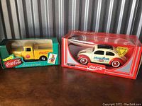 Photo of two vintage Coca-Cola metal die-cast toy vehicles in original packaging showing the 1956 delivery truck and the 1958 VW Coccinelle Beetle