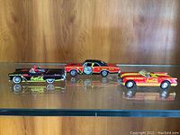 Three Coca-Cola themed Dinky model cars displayed side by side on a clear glass shelf against a wood background.