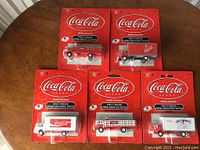 Set of five Ford C Series Coca-Cola delivery truck diecast models in original sealed packaging, arranged on a wooden table.