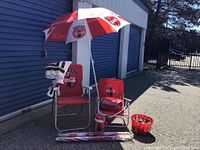 Two Coca-Cola folding lawn chairs, one with cushion, three Coca-Cola branded umbrellas, red bucket with glass collectible bottles, and red cooler jug arranged outside.