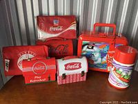 Photo showing 7 Coca-Cola themed lunch-related items arranged on a wooden surface: 2 tin lunchboxes, 2 collapsible lunch bags, 1 metal lunchbox with polar bear artwork, and 2 thermoses.