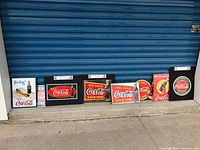 Seven used metal Coca-Cola signs arranged side by side, showing various sizes and vintage Coca-Cola logos and slogans.