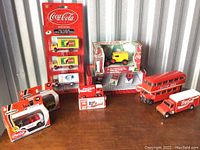 Photo showing several Coca-Cola branded vehicle models on a wooden surface including a double-decker bus, delivery trucks, and boxed items.