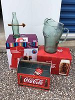Nostalgia Coca-Cola red ice maker, green embossed glass Coca-Cola pitcher, and metal Coca-Cola bottle carrier with wooden handle displayed together.