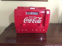Front view of red Coca-Cola cooler radio with tuning dial and knobs, showing Coca-Cola logo and 'ICE COLD' text.