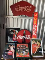 Group photo showing multiple Coca-Cola vintage signs including large red rivet logo sign, round sign, vertical 'ICE COLD Coca-Cola' sign, and various rectangular advertising signs with vintage artwork.