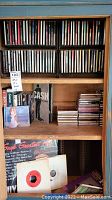 Wooden cabinet filled with various music CDs on shelves, a few smaller CDs stacked, some vinyl records at bottom.