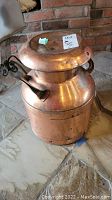 Full view of copper milk can standing on stone floor, showing overall shape, lid, body and handles.